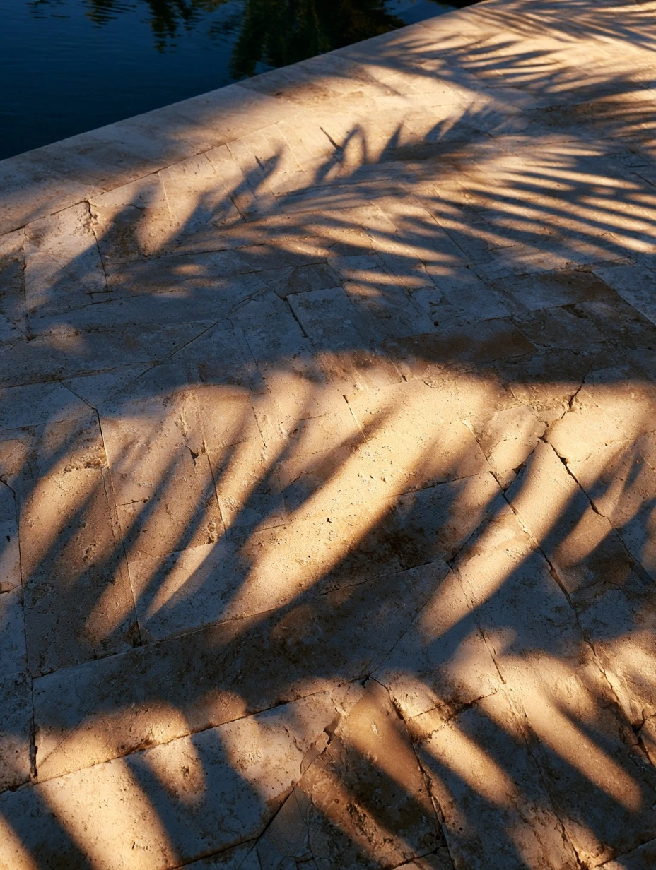 A pale stone pool deck running to the water edge under late afternoon palm shadows